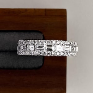 May include: A silver ring with a band of rectangular and round-cut gemstones. The ring is displayed on a dark wooden surface with a gray velvet lining. The gemstones sparkle, reflecting light.