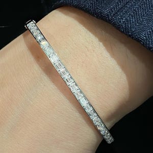 Op de afbeelding: Een delicate, zilverkleurige armband versierd met rechthoekig geslepen, heldere edelstenen. De stenen zijn in een lineair patroon geplaatst, waardoor een continu sprankelen ontstaat. De sieraden worden getoond op een pols, tegen een donkere achtergrond.