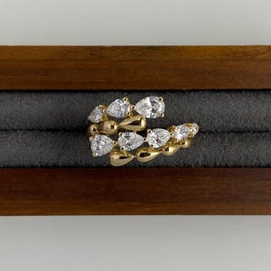 May include: A gold ring with a distinctive design, set with multiple clear, pear-shaped gemstones. The ring is displayed in a wooden jewellery box with a grey interior. The ring's design curves around the finger, showcasing the gemstones.
