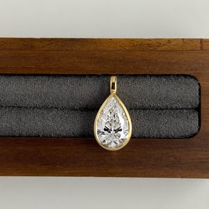 May include: A pear-shaped clear gemstone pendant set in a polished yellow gold bezel. The pendant features a single faceted stone with a pointed top and rounded base, secured by a small gold bail at the apex, showcasing a brilliant reflective surface.