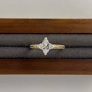 May include: A gold ring with a four-petal flower-shaped diamond center stone. The band is also adorned with smaller diamonds. The ring is displayed in a dark wooden jewelry box with a gray velvet interior.