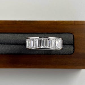 May include: A white gold ring featuring a continuous band of rectangular-cut gemstones. The ring is presented in a wooden jewellery box with a dark grey interior. The gemstones are clear and reflect light.