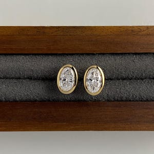 Könnte beinhalten: Ein Paar goldene, oval geformte Ohrstecker mit einem diamantartigen Mittelteil. Die Ohrringe sind in einer goldenen Lünette gefasst und liegen auf einer grauen Samtoberfläche in einer hölzernen Schmuckschatulle.