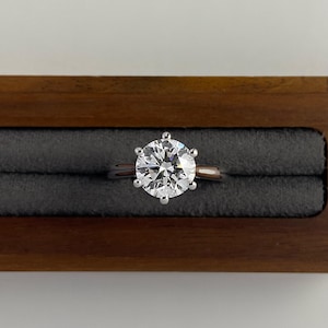 May include: A solitaire engagement ring with a large, round, brilliant-cut diamond set in a silver band. The ring is displayed in a dark wooden ring box with a gray velvet interior. The diamond sparkles brightly.