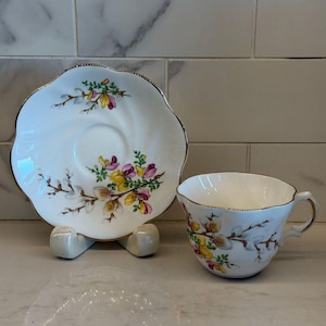 May include: A white teacup and saucer set with a floral design in yellow, pink, and green. The edges of the cup and saucer are scalloped and trimmed with gold. The saucer is propped up on a white stand.