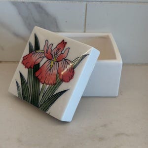 May include: A square white ceramic trinket box with a removable lid. The lid features a hand-painted illustration of a pink iris flower with green leaves. The box has a smooth, glossy finish and a simple, clean design for storing small items.