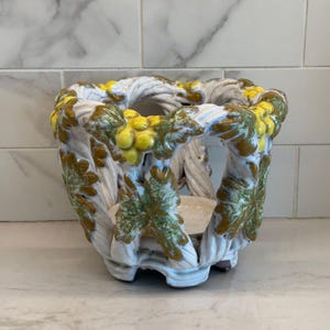 May include: A white ceramic planter with a decorative design of yellow grapes and green leaves. The planter has a woven texture and a round opening. The base is a light beige color.