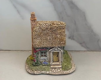 Może przedstawiać: Miniaturowa figurka domku z trzcinowym dachem, ceglanym kominem i małymi drzwiami. Domek otoczony jest zielenią i kwiatami, z kamienną podstawą. Paleta kolorów obejmuje beż, brąz, zieleń i akcenty czerwieni i błękitu.