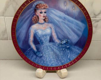 May include: A decorative plate featuring a bride in a blue gown and veil. The plate has a maroon border with gold accents. The bride is holding a bouquet. The plate is supported by a white stand.