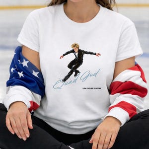 May include: White t-shirt featuring a figure skater in a black outfit, with the text "Quad God" and "USA FIGURE SKATERS". The person is wearing black pants, white ice skates, and a jacket with an American flag design.