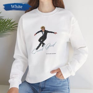 Puede incluir: Sudadera blanca de cuello redondo con un gráfico de un patinador artístico con un atuendo negro. El texto "Quad God" y "USA FIGURE SKATERS" están impresos debajo. La palabra "White" está en una pancarta azul en la esquina superior izquierda.