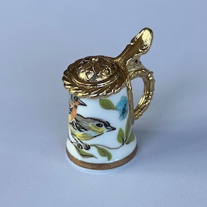 Puede incluir: Una jarra de cerámica blanca en miniatura con asa, tapa y ribete dorados. La jarra está decorada con pájaros pintados a mano, flores azules y hojas verdes. La jarra es un objeto de colección decorativo.