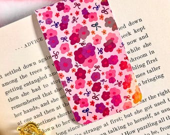 May include: A floral patterned bookmark in shades of pink and purple is inserted into a book. The bookmark features a repeating pattern of flowers and bows. A white pen with a gold crown-shaped top rests on the open book.