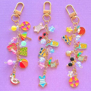 May include: Three gold-toned metal keychains featuring assorted summer-themed charms including swimsuits, sunglasses, beach balls, umbrellas, and pool floats. Each keychain is attached to a lobster clasp and decorated with colourful beads and charms.