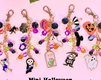 May include: A collection of gold-toned metal bag charms featuring Halloween-themed enamel pendants including ghosts, pumpkins, skeletons, and tombstones with the text RIP, accented with colorful beads and lobster clasps on a pink background.