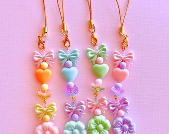 May include: Four beaded phone charms featuring pastel-colored plastic bows, hearts, and flower-shaped beads. Each charm is attached to a gold-toned metal lobster clasp and a thin gold cord, finished with a small gold-toned metal star charm at the bottom.