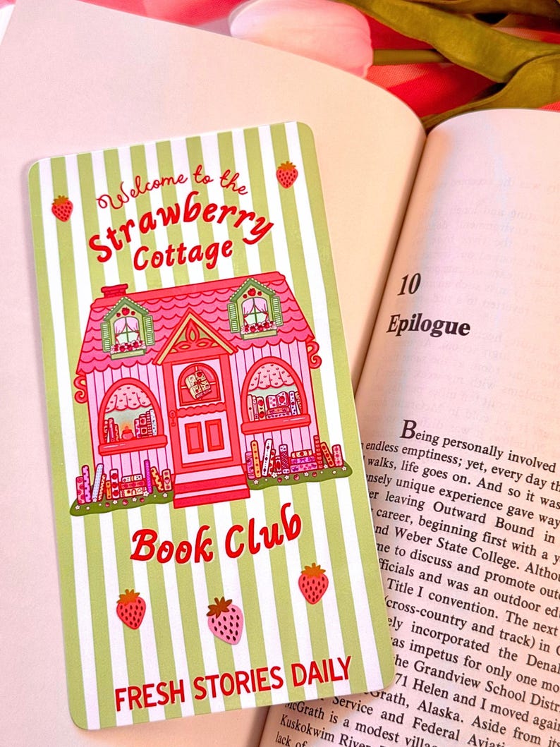 A pastel-colored wide bookmark with a pink cottage design sits on an open book. The card reads "Welcome to the Strawberry Cottage Book Club" and features strawberries and green stripes, creating a whimsical and inviting feel.