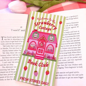 A pastel-colored wide bookmark with a pink cottage design sits on an open book. The card reads "Welcome to the Strawberry Cottage Book Club" and features strawberries and green stripes, creating a whimsical and inviting feel. In the background, a tulip and a checkered cloth add to the cozy atmosphere.