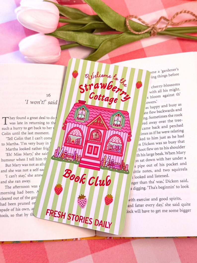 A pastel-colored wide bookmark with a pink cottage design sits on an open book. The card reads "Welcome to the Strawberry Cottage Book Club" and features strawberries and green stripes, creating a whimsical and inviting feel. In the background, a tulip and a checkered cloth add to the cozy atmosphere.
