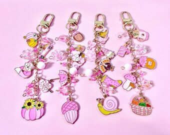 May include: Four gold-toned metal keychains featuring assorted charms including pink pumpkins, sunflowers, acorns, snails, mushrooms, and baskets. Each keychain is decorated with translucent pink beads, white spherical beads, and gold-toned lobster clasps.