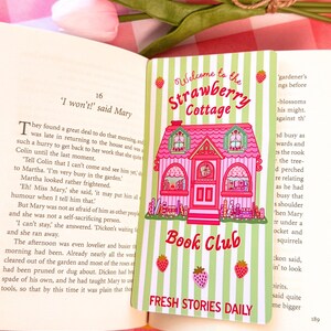A pastel-colored wide bookmark with a pink cottage design sits on an open book. The card reads "Welcome to the Strawberry Cottage Book Club" and features strawberries and green stripes, creating a whimsical and inviting feel. In the background, a tulip and a checkered cloth add to the cozy atmosphere.