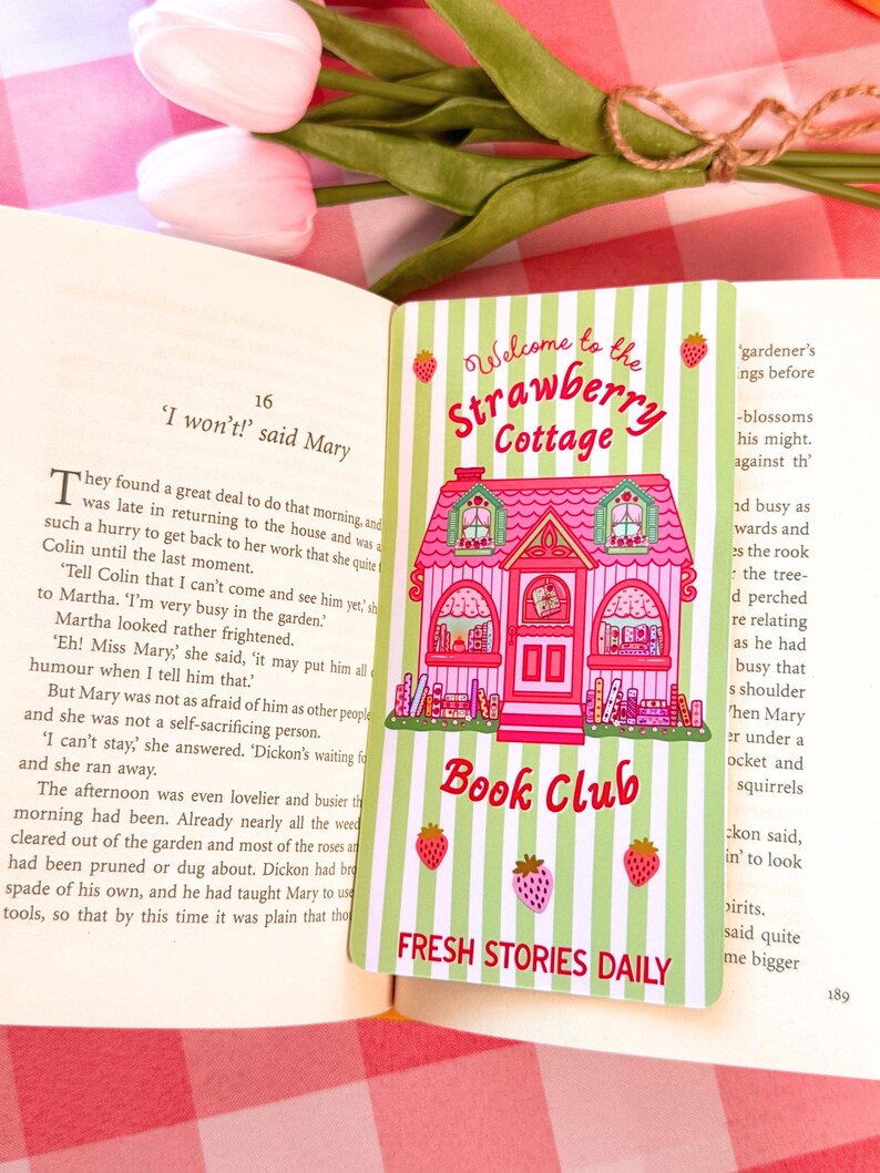 A pastel-colored wide bookmark with a pink cottage design sits on an open book. The card reads "Welcome to the Strawberry Cottage Book Club" and features strawberries and green stripes, creating a whimsical and inviting feel. In the background, a tulip and a checkered cloth add to the cozy atmosphere.