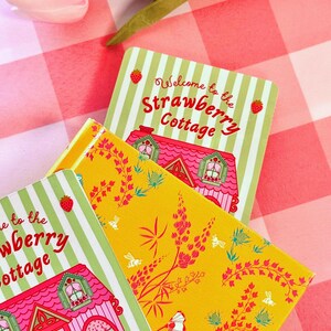 A colorful scene with pink tulips on a checkered tablecloth. A vibrant yellow floral-patterned book lies beneath a hand drawn "Strawberry Cottage" bookmark. Bright and cheerful.
