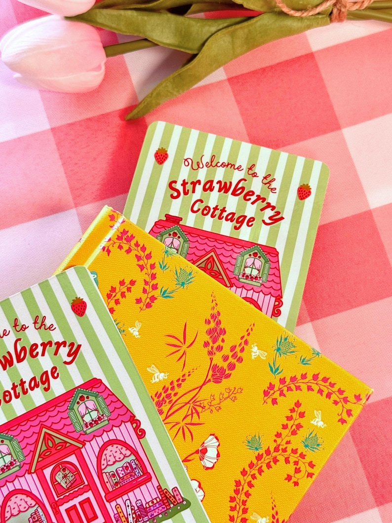 A colorful scene with pink tulips on a checkered tablecloth. A vibrant yellow floral-patterned book lies beneath a hand drawn "Strawberry Cottage" bookmark. Bright and cheerful.