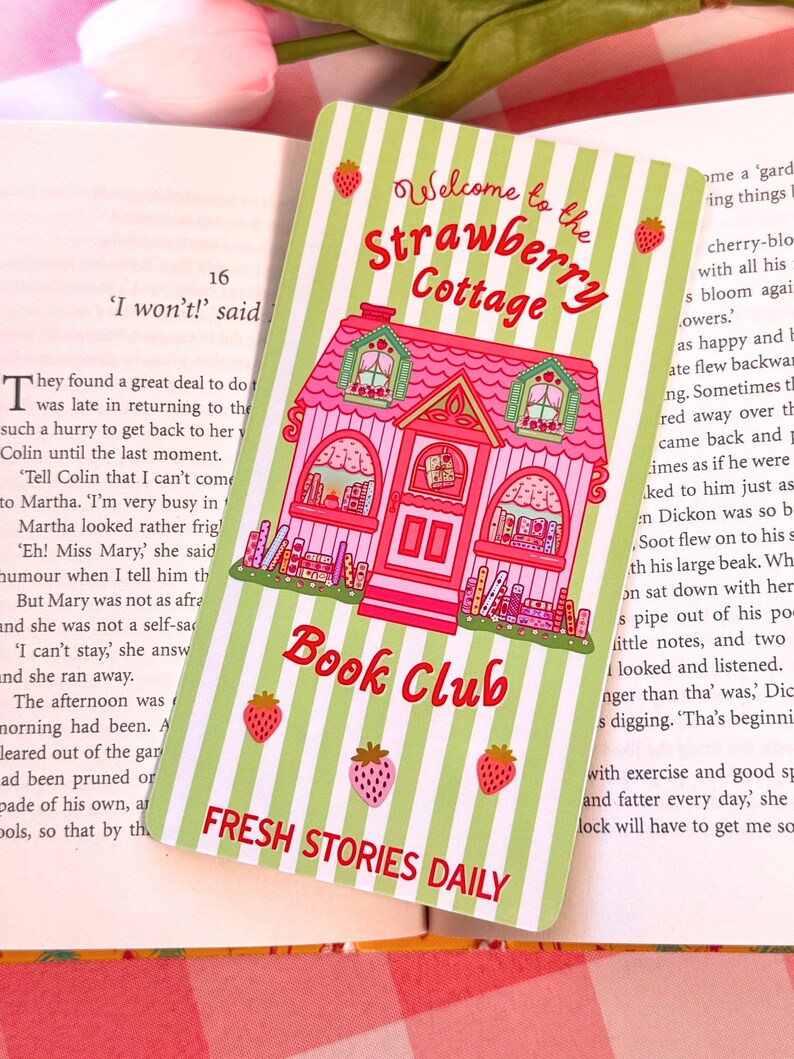 another shot of a pastel-colored wide bookmark with a pink cottage design sits on an open book. The card reads "Welcome to the Strawberry Cottage Book Club, Fresh Stories Daily" and features strawberries and green stripes, creating a whimsical and inviting feel.