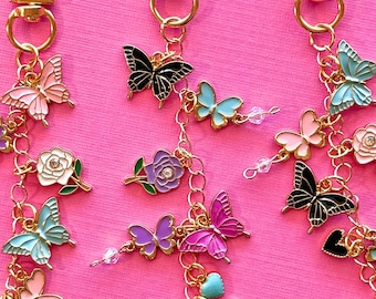May include: Three gold-toned metal keychains featuring dangling charms of colorful butterflies, flowers, and hearts. Each keychain has a lobster clasp and includes small clear beads. The charms are finished in enamel colors including pink, blue, purple, black, and white.