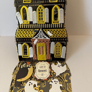 May include: A three-dimensional paper craft haunted house in black and yellow with glittery accents. The structure features arched windows, shutters, and ghosts. A front panel displays an owl, two ghosts, and a sign reading IF YOU GOT IT haunt IT.