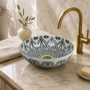 Puede incluir: Un lavabo de sobreponer redondo con un patrón geométrico complejo en blanco y negro tanto en el interior como en el exterior, junto con un grifo de metal dorado y un desagüe a juego también dorado en el centro.
