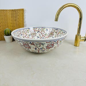 Peut inclure: Une vasque ronde en céramique ornée d'un motif floral peint à la main avec des vignes roses, vertes et bleues sur fond blanc. Le lavabo est placé à côté d'un robinet en laiton courbé et d'un petit cactus en pot dans un contenant blanc.