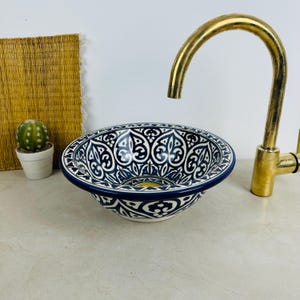 Może przedstawiać: Okrągła ceramiczna umywalka nablatowa z misternym geometrycznym wzorem w kolorze ciemnoniebieskim i białym. Misa znajduje się obok wysokiej, zakrzywionej mosiężnej baterii. Mały zielony kaktus w białej doniczce stoi po lewej stronie umywalki.