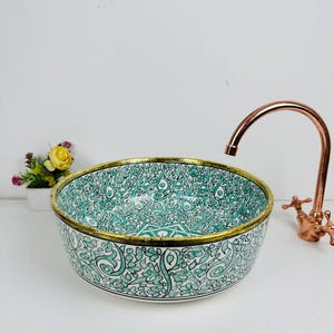 Puede incluir: Un lavabo de cerámica redondo con un patrón floral detallado en turquesa y negro en sus superficies interior y exterior, acabado con un borde dorado metálico y acompañado de un grifo curvo de tono cobre.