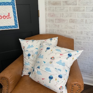 May include: Two white throw pillows with a blue and white hot air balloon pattern on a brown leather armchair. The pillows have a pocket on the back.