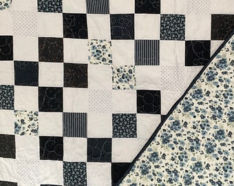 May include: A white quilt with a black and white checkered pattern. The quilt is made up of different fabric squares with various patterns, including floral, polka dot, and striped. The back of the quilt is a floral print.