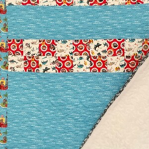 May include: A blue and red patchwork quilt with a white and blue wave pattern on the back. The quilt features a repeating pattern of pirates, sharks, and treasure chests.