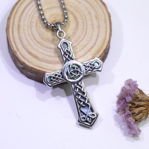 May include: A silver-toned Celtic cross pendant necklace. The cross features intricate knotwork and a central Trinity knot design. The pendant has iridescent accents and hangs from a matching chain. A small bundle of dried purple flowers rests nearby.