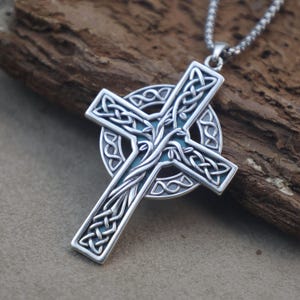 May include: A silver-toned metal pendant in the shape of a Celtic cross, featuring intricate knotwork patterns and a central vine-like design. The cross is layered over a circular ring and hangs from a silver-toned metal ball chain necklace.