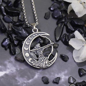 May include: A silver-colored pendant necklace featuring a crescent moon design with an archer figure. The necklace hangs on a chain, set against a backdrop of black stones and white flowers. The pendant is approximately 1.5 inches in diameter.