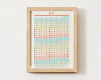 May include: A framed 2026 yearly calendar with a light wood frame. The calendar grid is divided into pastel colors, with the year "2026" in orange at the top. The calendar is displayed on a white wall.