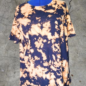 Puede incluir: Una camiseta azul marino con un patrón de tinte de lejía en tonos melocotón y crema. La camiseta de manga corta se muestra en un maniquí azul sobre un fondo gris texturizado. La camiseta tiene cuello redondo.