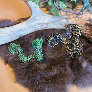 以下が含まれることがあります： 緑、黒、金色のワイヤーアート彫刻が3つ。1つは様式化されたヘビ、もう1つはスパイラル、3つ目は背の高い円筒形です。彫刻は茶色の毛皮のような表面に展示されています。