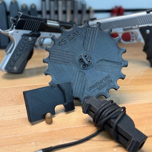 Könnte beinhalten: Ein schwarzes, 3D-gedrucktes, zahnradförmiges Gerät mit dem Text "45 CAL", ein schwarzes rechteckiges Teil und ein schwarzes Gerät mit einem Kabel. Eine Kugel und zwei silberne und schwarze Handfeuerwaffen sind ebenfalls sichtbar.