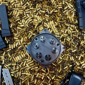 Könnte beinhalten: Draufsicht auf eine Ansammlung von Messing-Patronenhülsen. Ein metallisch graues Objekt mit kreisförmigen Merkmalen befindet sich in der Mitte. Schwarze Waffenmagazine und ein kleines schwarzes Lederetui deuten auf ein Thema im Zusammenhang mit Schusswaffen und Munition hin.