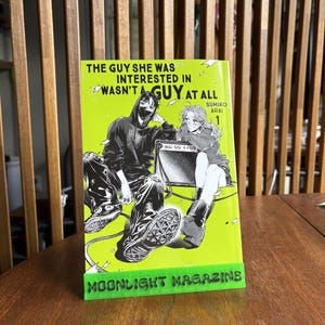 May include: A manga book titled "THE GUY SHE WAS INTERESTED IN WASN'T A GUY AT ALL" with a neon green cover and a black and white illustration of two people. The book is propped up on a green stand that reads "MOONLIGHT MAGAZINE".