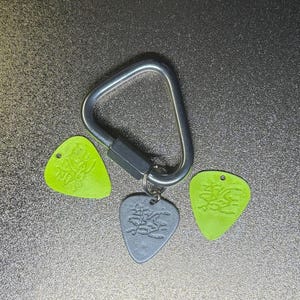 Op de afbeelding: Een zilverkleurige metalen karabijnhaak met drie plectrums. Twee groene plectrums en één grijze plectrum zijn bevestigd. Elk plectrum heeft een ontwerp in het oppervlak geëtst. De plectrums hebben een driehoekige vorm.