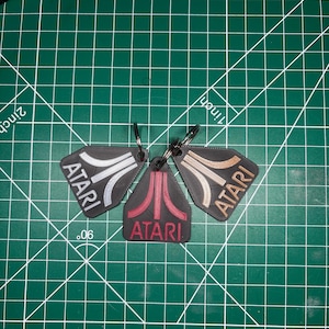 Puede incluir: Tres llaveros de plástico negro con el logotipo y el texto de Atari. Un llavero tiene el logotipo blanco, otro rojo y otro de color cobre. Cada llavero está unido a una anilla de metal y mide aproximadamente 3,8 cm de ancho.