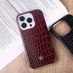 Puede incluir: Una funda de teléfono burdeos con textura de piel de cocodrilo y una letra dorada "W" se muestra junto a una funda de teléfono negra con una textura similar. Las fundas están sobre una superficie de madera, con un libro abierto al fondo.